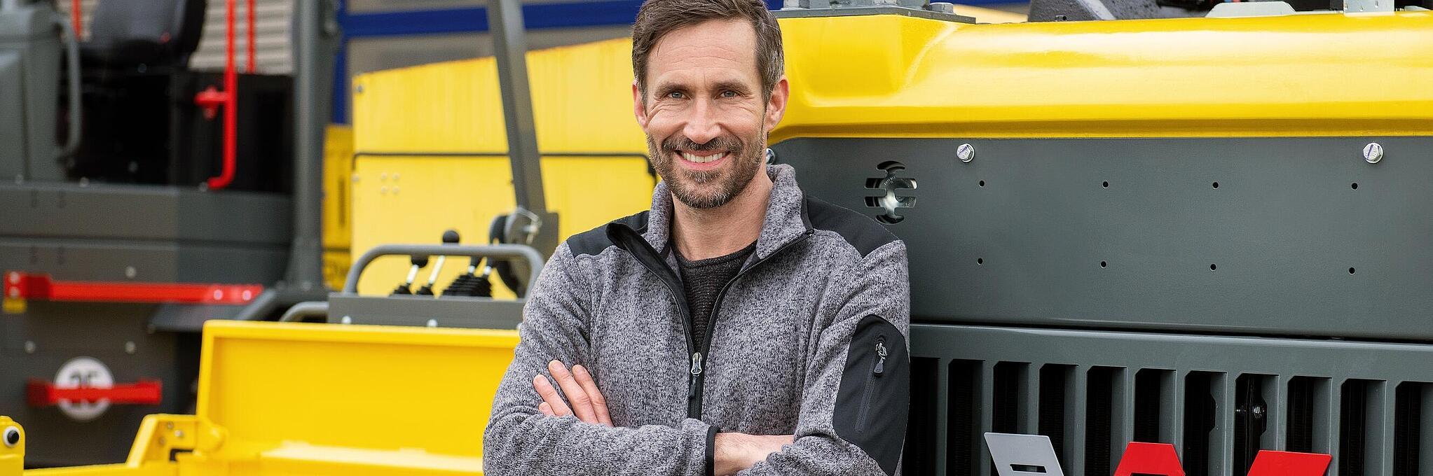 Smiling Wacker Neuson employee, arms crossed, leaning against a Wacker Neuson construction machine.